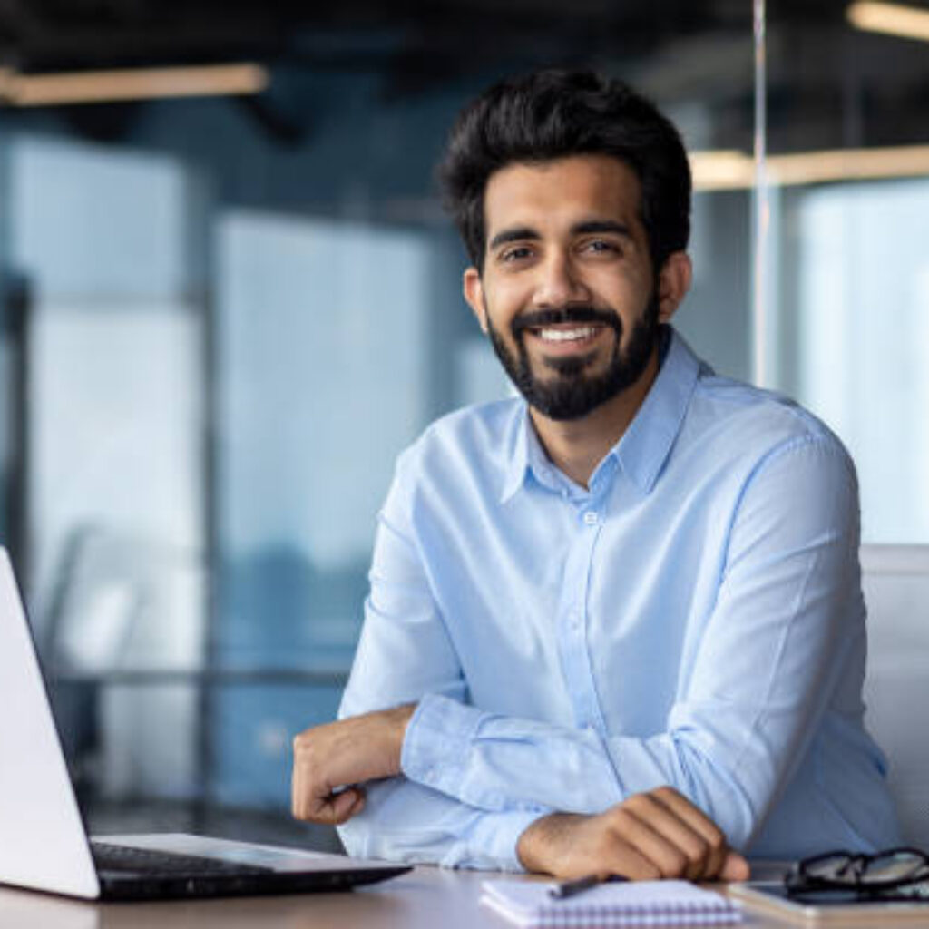 Portrait of happy and successful businessman, indian man smiling and looking at camera, satisfied with achievement results man working inside office building using laptop at work.